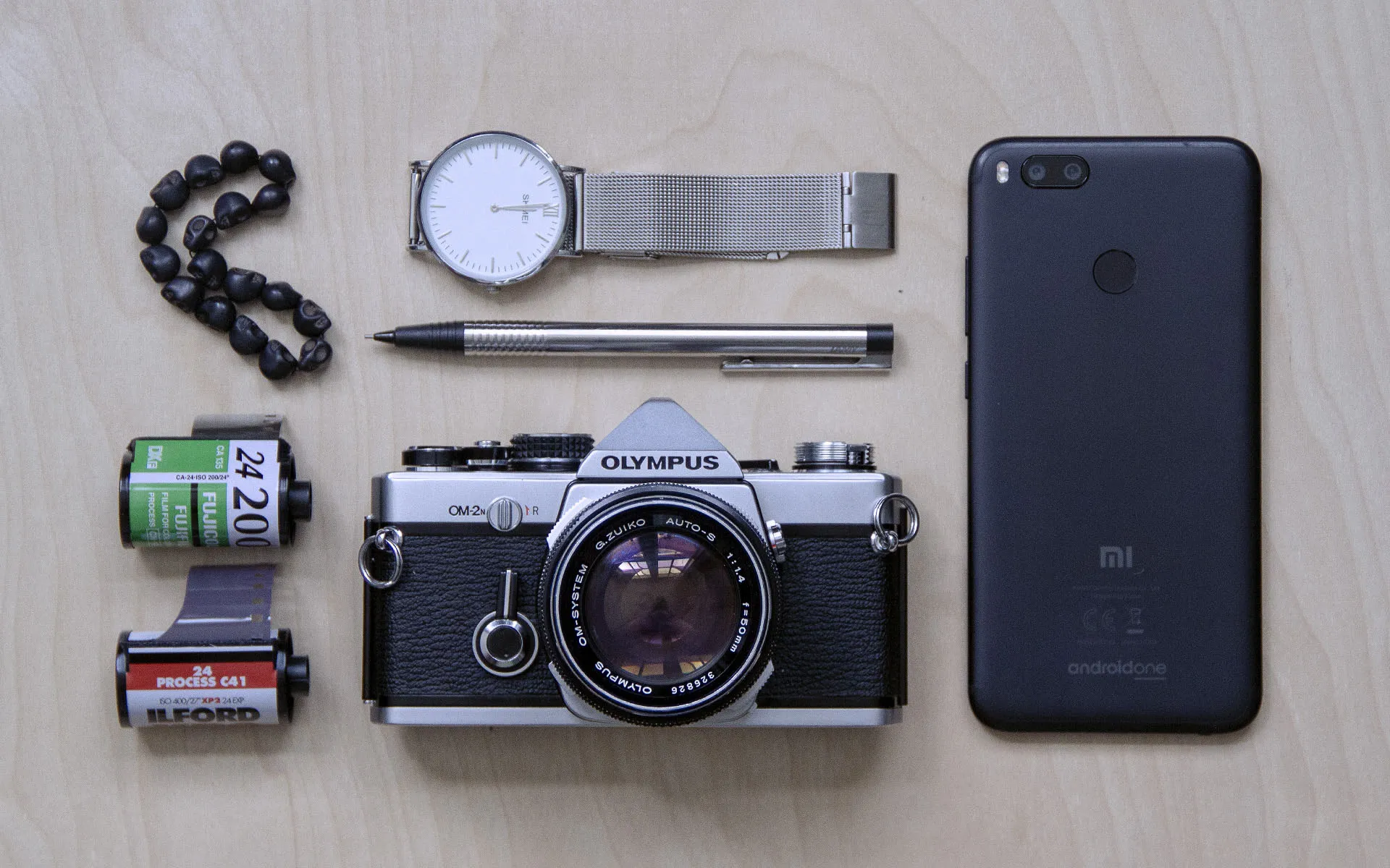 Olympus OM-2N, two rolls of film, a watch, a bracelet, mechanical pencil and a smartphone knolled on the table.