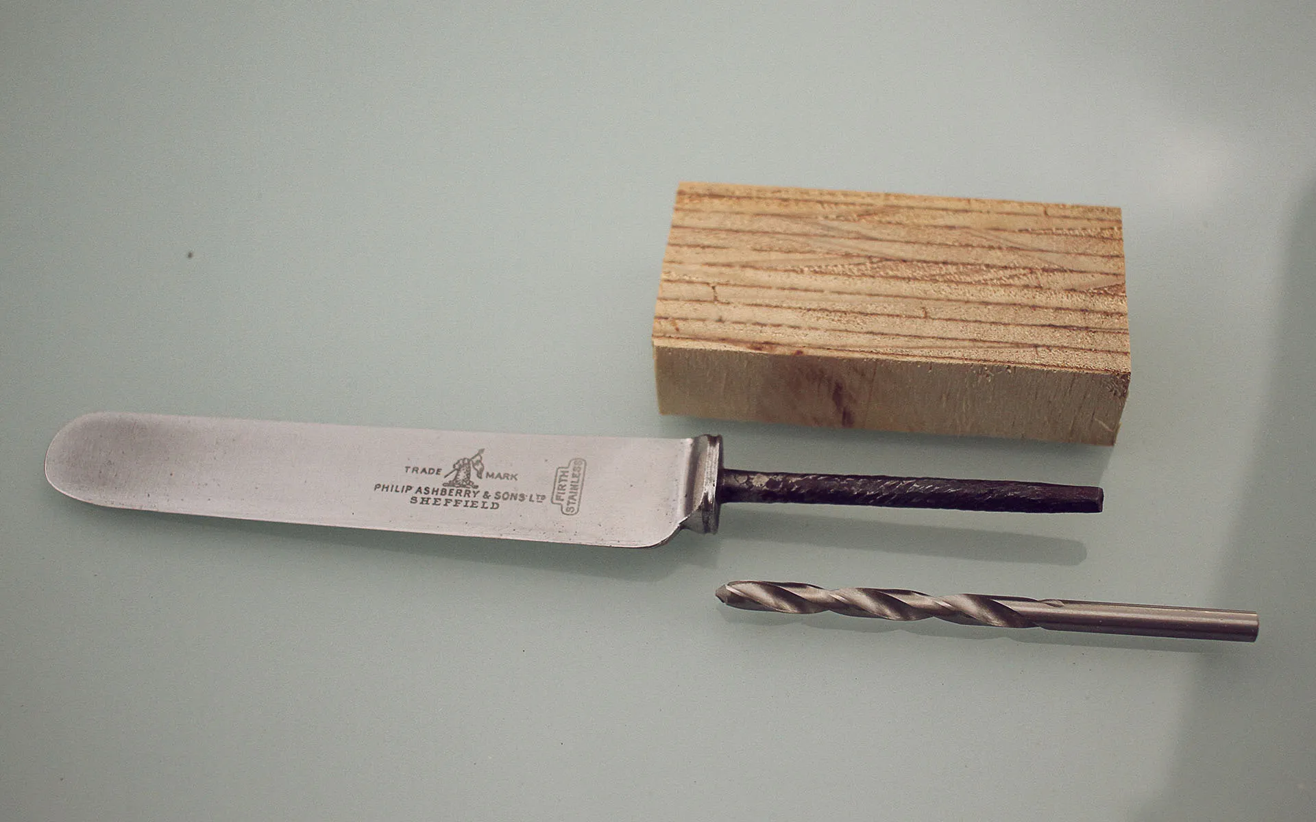 Knife blade without handle next to a block of wood and a drill bit.